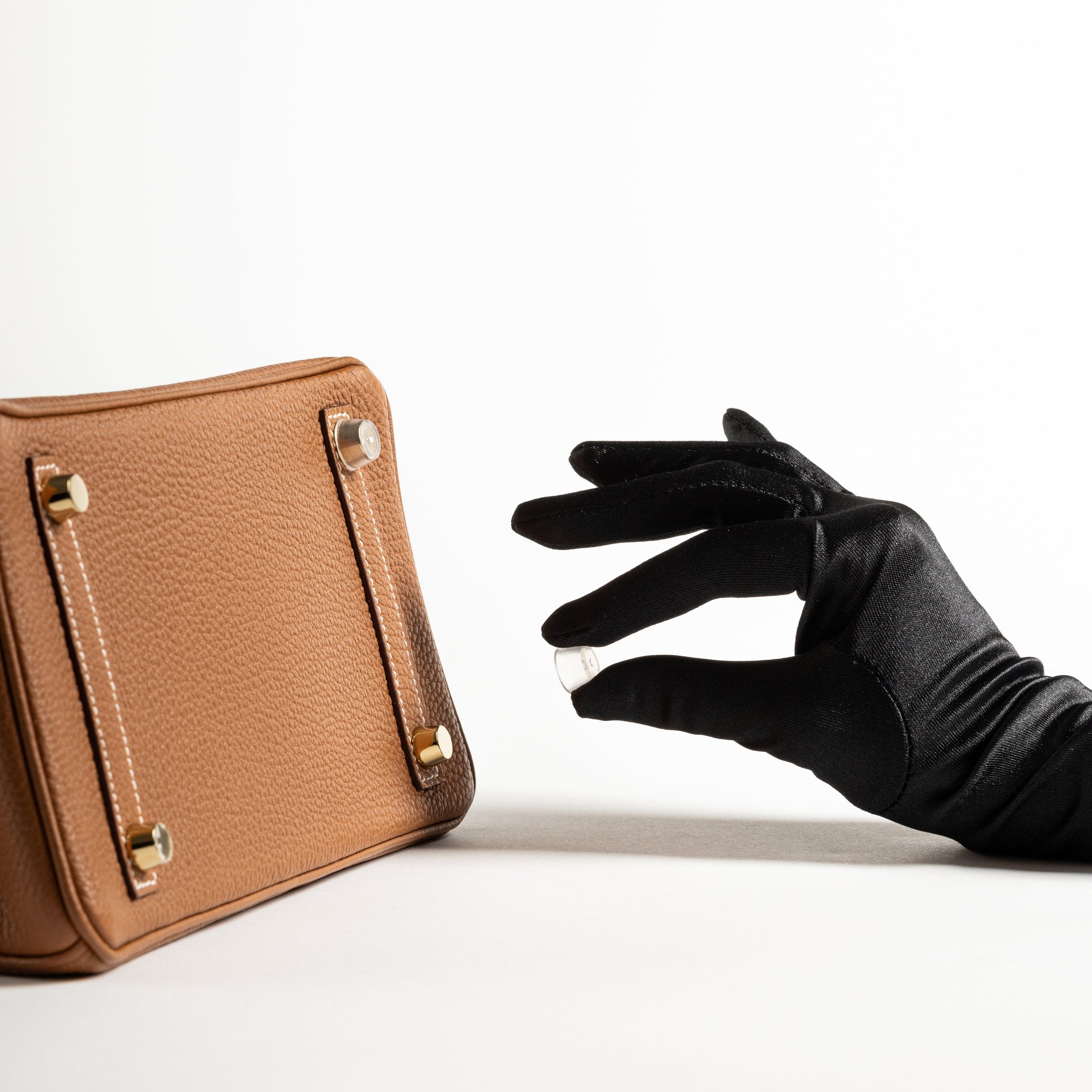 Brown leather handbag with a black gloved hand reaching towards it on a white background