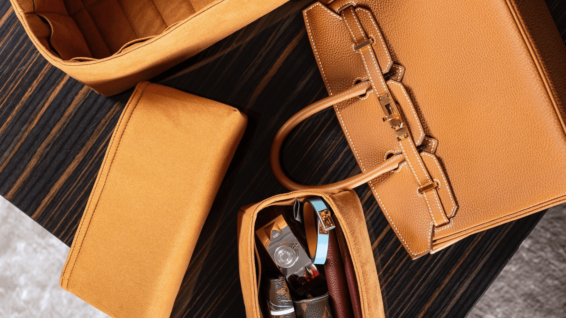 A tan leather handbag organizer set including a main compartment, a zippered pouch, and a slim wallet, arranged on a dark wood surface next to a structured tan handbag.